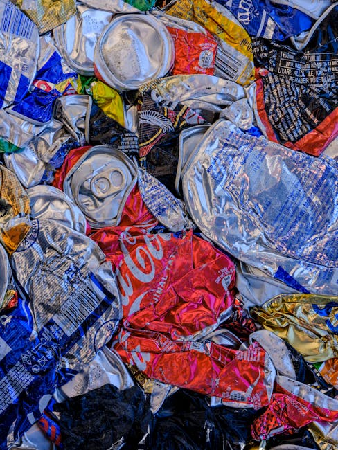 A close-up image of a tightly packed pile of crushed aluminum cans, predominantly in silver with some displaying partial branding in red, blue, yellow, and black. The cans are varied in size and shape, with some showing visible pull tabs and others with torn or flattened tops, indicating previous use and disposal. The surface of the cans is shiny and metallic, reflecting ambient light, amidst a mixture of crumpled, wrinkled, and layered packaging. The cans are situated outdoors on a flat surface, possibly on a pavement or driveway, with no additional context visible such as waste containers or surrounding environment. This scene represents waste collection of beverage containers, highlighting the importance of proper disposal and the potential for recycling, which is relevant to professional rubbish removal services such as those offered by Rubbish Collection Bow, particularly when considering alternative waste handling methods beyond regular council collections.