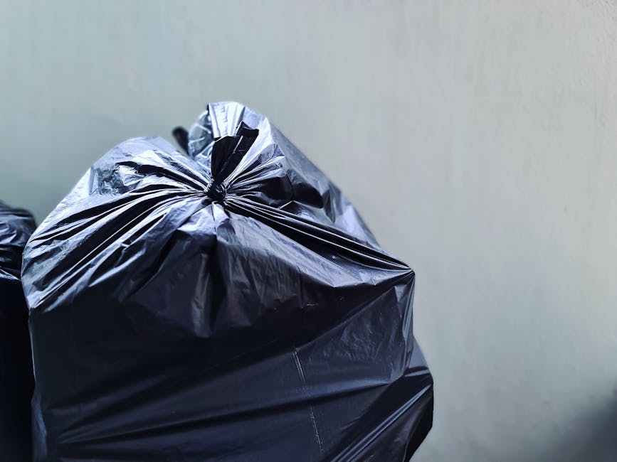Close-up of a large black plastic rubbish bag filled with waste, tightly knotted at the top, placed outdoors against a plain light-colored wall. The bag exhibits a shiny, crinkled texture with visible creases and folds, indicating it is full and heavy. Nearby, additional black rubbish bags are partially visible, suggesting a collection of waste for removal. The setting appears to be on a pavement or driveway, typical of residential or commercial exterior disposal areas. The lighting is natural, casting soft shadows on the bag’s surface. This scene exemplifies an independent rubbish disposal method, often managed by private waste collection services such as Rubbish Collection Bow, providing an alternative to traditional council bin collections for efficient waste clearance.