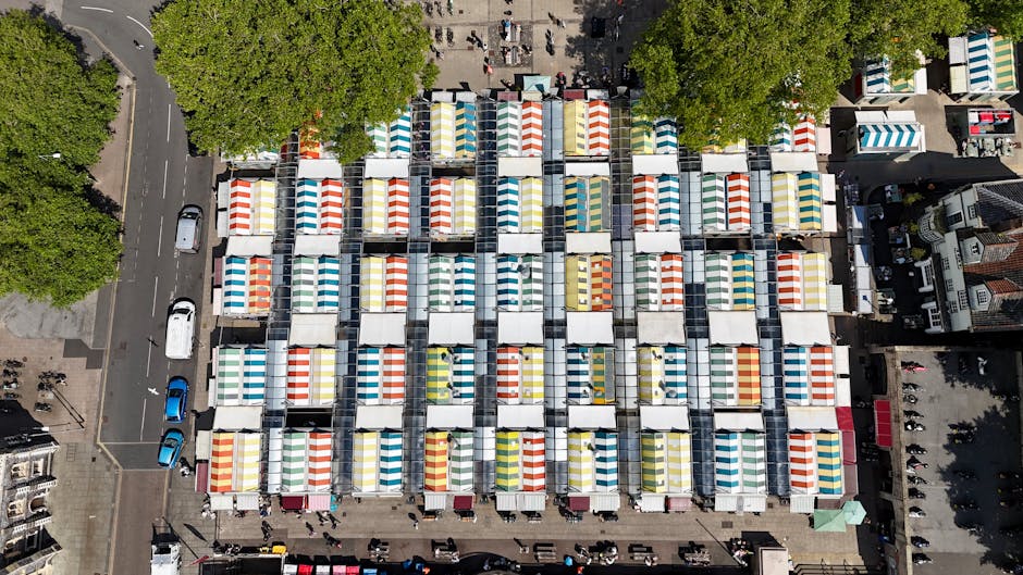 An aerial view of a bustling market square populated with numerous market stalls, each covered with colorful striped canopies in shades of red, yellow, blue, green, and white. The stalls are arranged in a grid pattern, with narrow pathways separating them, and are situated on a paved surface. Surrounding the market are several large, leafy green trees providing shade, along with adjacent streets lined with parked cars and moving vehicles. To the right and bottom edges of the image, residential buildings with brick facades and pitched roofs are visible, framing the square. The scene is illuminated with natural daylight, creating a bright and lively atmosphere typical of an outdoor marketplace during daytime. In the context of waste management, this image showcases an organized market environment where companies such as Rubbish Collection Bow might provide scheduled rubbish removal or private waste disposal services for market vendors and local businesses, highlighting an example of on-site clearance in a commercial setting.