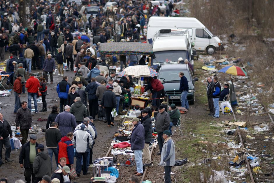 A large outdoor market scene shows numerous vendors and shoppers on a muddy, uneven ground with scattered rubbish and debris along the sides. In the foreground, several tables and makeshift stalls display various goods, with some items covered by umbrellas or shaded by small awnings. People are gathered around, browsing and interacting, dressed in casual jackets and hats suitable for cool weather. Behind the stalls, a line of parked vehicles, including white vans and small trucks, are positioned along a dirt pathway, suggesting a temporary or mobile market setup. The background features more shoppers extending into a densely packed area, with trees and a slightly overcast sky overhead. The ground and surrounding area are cluttered with discarded packaging, plastic bags, and other waste materials, illustrating the environmental impact often linked with outdoor markets and street trading. Rubbish collection and waste management are implied by the presence of rubbish scattered amid the activity, highlighting the importance of proper waste disposal services like those provided by Rubbish Collection Bow for alternative waste handling and clearance solutions.