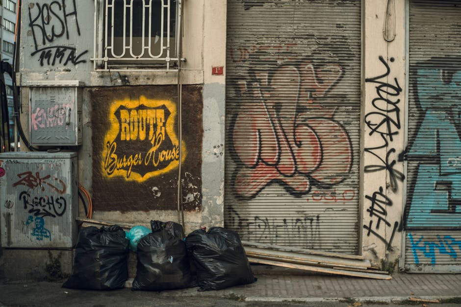 An urban street scene featuring a graffiti-covered building façade with a closed metal roller shutter door painted in shades of tan and rust, displaying large, stylized graffiti tags in black, red, and blue. To the left, a small window with white bars is partially visible above a dark brown wall that has a yellow painted sign reading 'Rough Burger House' with black lettering. In front of the building, on a paved sidewalk, there are four tightlypacked black rubbish bags, possibly filled with waste or discarded items, positioned at the base of the wall. Nearby, an electrical box with graffiti tags and some exposed cables is mounted on the wall. The overall scene highlights typical elements associated with city waste and debris, with the rubbish bags as the primary focus relating to rubbish removal services and private waste handling.