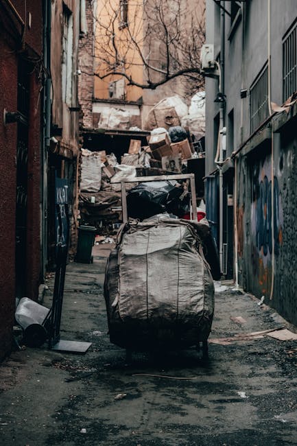 A narrow urban alleyway cluttered with piled rubbish and discarded materials, with a large, wheeled trash bag prominently positioned in the foreground. The bag appears to be made of heavy-duty, weathered fabric or plastic, and is partly filled with waste, showing a crumpled texture. Surrounding the bag, the asphalt ground is stained and uneven, scattered with smaller debris and partially obscured trash items. On either side, there are buildings with exposed brick and metal fencing; the left side features a red wall with graffiti, while the right side has a gray, painted exterior with barred windows. In the background, a dense heap of cardboard boxes, black plastic bags, and miscellaneous waste materials is stacked against a structure, indicating an area designated for rubbish collection or alternative waste handling. Overhead, leafless tree branches extend across the scene, contrasting the urban environment with natural elements, under overcast daylight that enhances the gritty, neglected atmosphere. This scene visually reflects the kind of refuse collection often managed by private waste removal services like Rubbish Collection Bow, highlighting the need for organized rubbish clearance in densely populated, space-constrained areas.