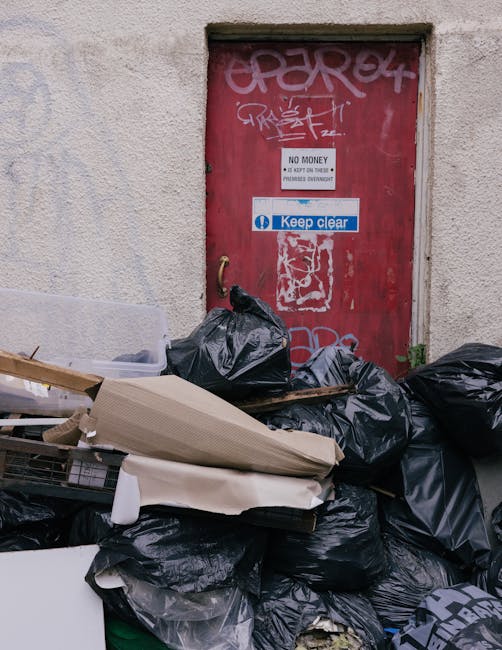 A close-up view of a red metal door set into a textured, off-white concrete wall. The door is covered with various graffiti tags and a white and blue 'Keep clear' sign, along with a smaller sign stating 'No money, do not open or tamper.' In front of the door, there is a large pile of discarded rubbish, primarily black plastic trash bags, some torn open, revealing rubbish inside. Among the debris are flattened cardboard boxes, a piece of white foam packaging, and a wooden pallet leaning against the pile. The rubbish and debris are spread across a concrete surface, indicating a collection or clearance scene typical of a private waste disposal area. The scene is lit naturally, emphasizing the clutter and the weathered appearance of the surroundings, which align with the context of waste management or alternative rubbish collection services such as those provided by Rubbish Collection Bow.