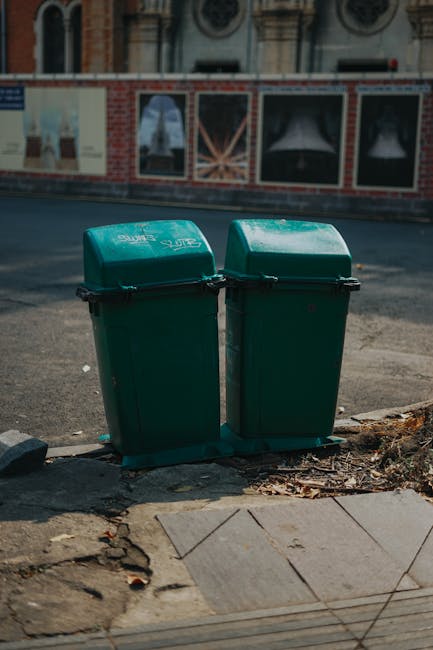 A row of six large black wheeled rubbish bins with closed lids is positioned on a pavement in front of a red brick wall. The bins have brightly coloured, predominantly orange and blue lids, with some lids showing orange on the left side and blue on the right side, all made of durable plastic material. The lids are flat with a slight texture, and the bins are uniformly aligned, suggesting organized waste collection. The environment appears to be outdoors, possibly along a residential or commercial building frontage, with natural daylight illuminating the scene. The arrangement of bins indicates organized waste or rubbish disposal, which is relevant to private rubbish removal services like those offered by Rubbish Collection Bow, providing an alternative to municipal waste collection in accordance with local guidelines for Bow waste management.